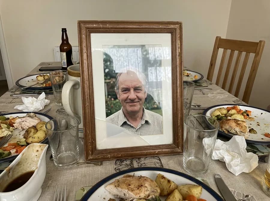 An old framed photograph of a man in his sixties propped on a family dinner table among half-finished roast dinners, with an empty chair pulled out behind it.