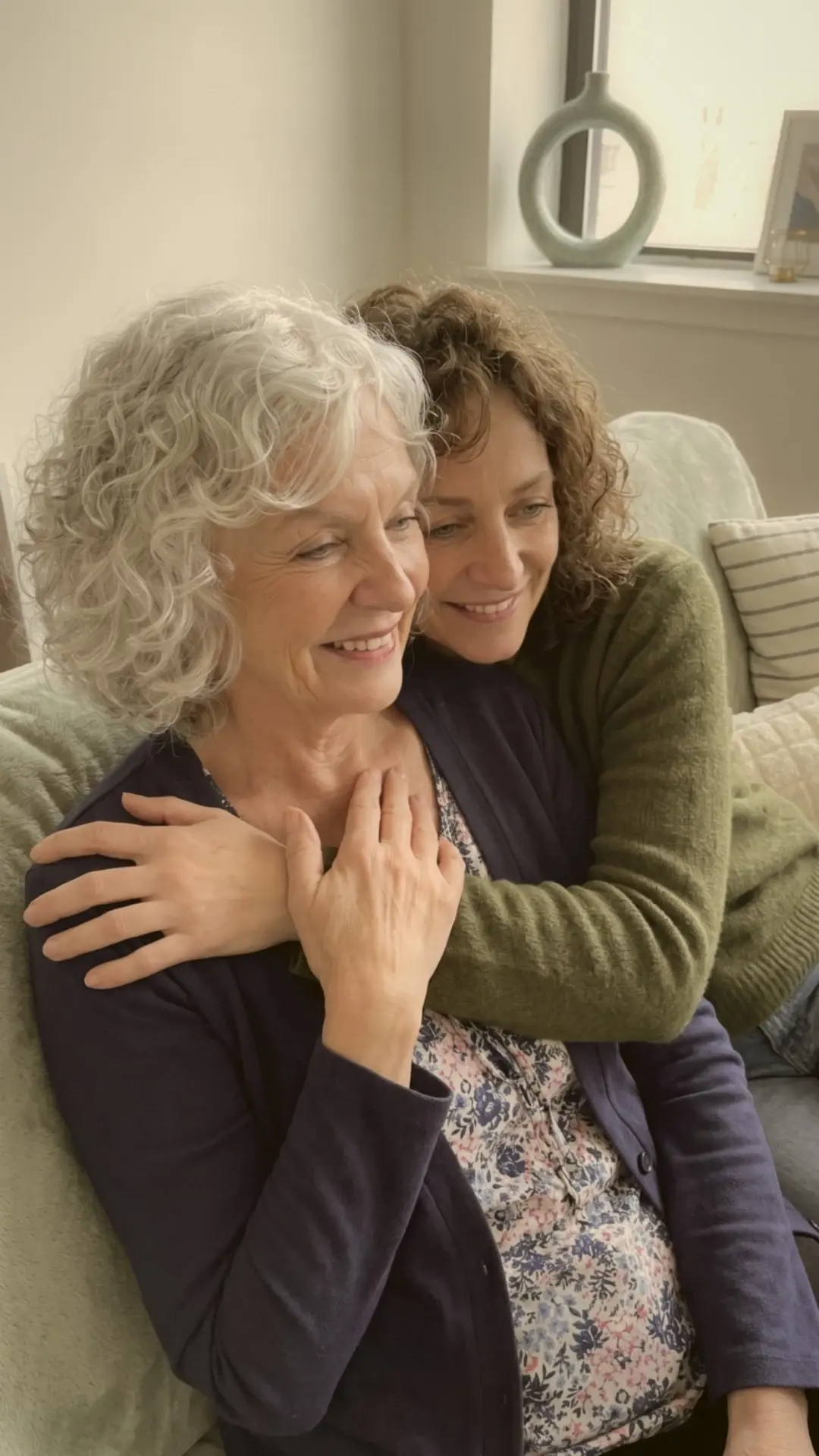 A woman in her sixties smiling with her hand on her heart, her grown daughter hugging her from behind on the sofa.