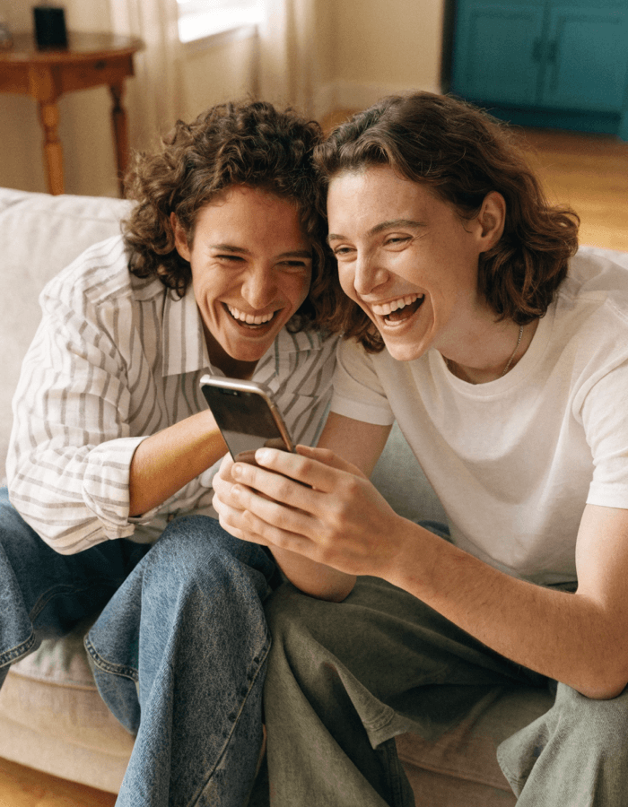 Two friends on a sofa laughing and wiping tears as they listen to a song on a phone between them.
