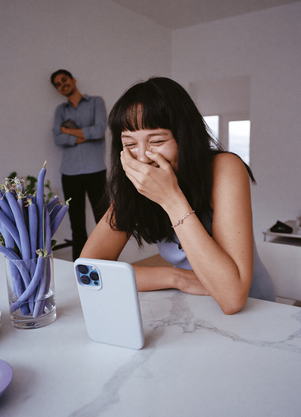 A woman at a white kitchen table, hand over her mouth, laughing through tears as a song plays from a phone in front of her.