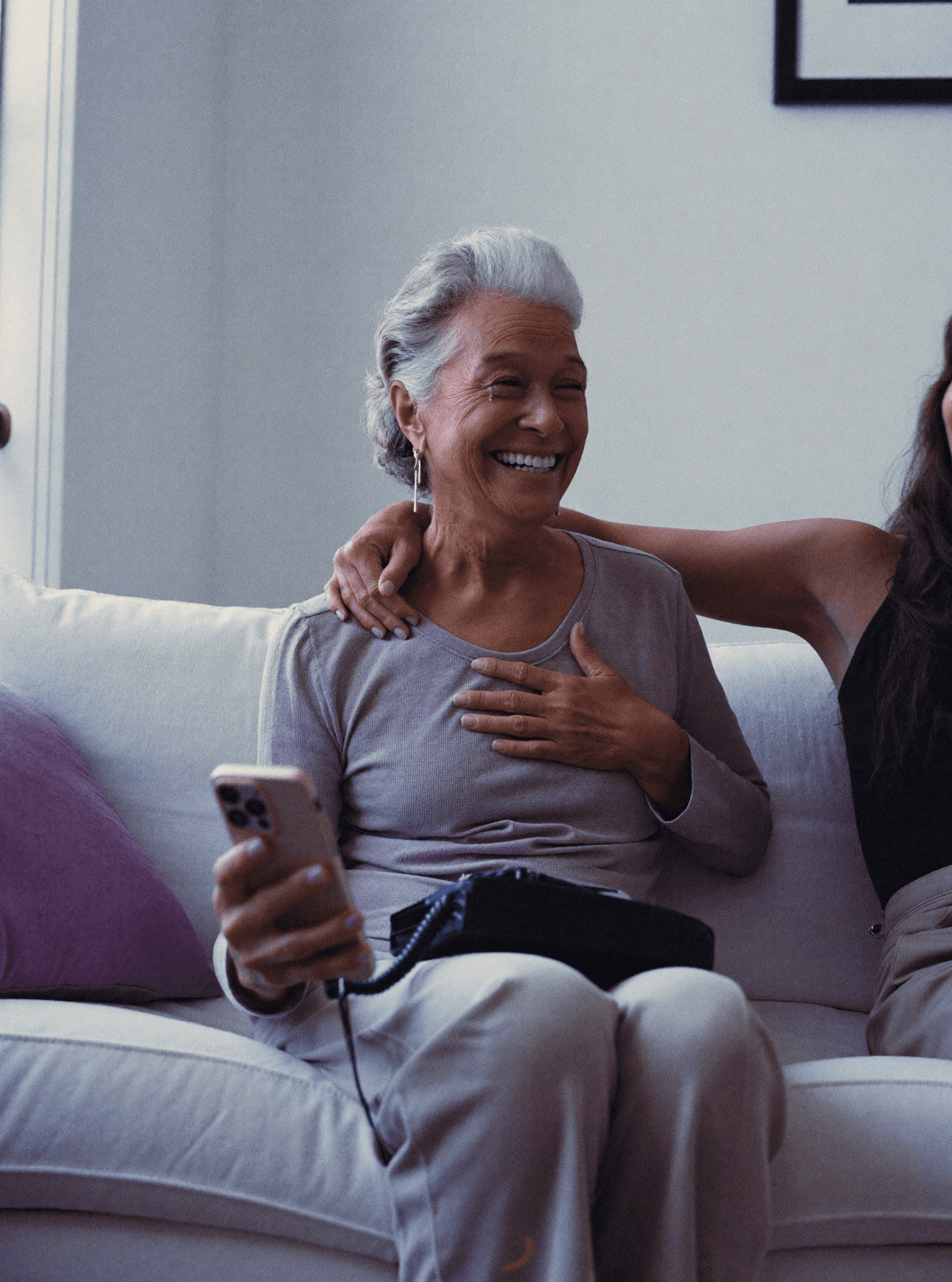 A woman in her late sixties holding a phone to her chest, smiling through tears, her daughter's arm around her.