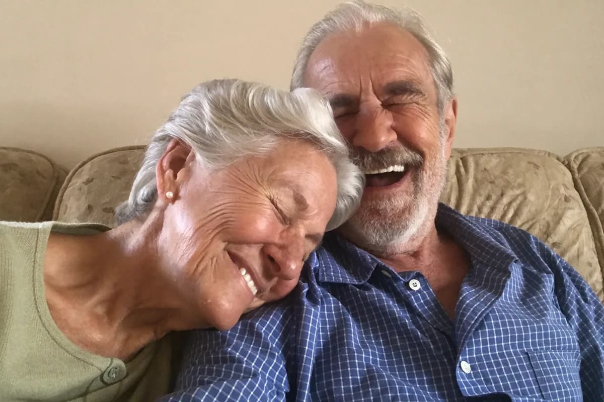 A couple with silver hair laughing together on a sofa at home, her head resting on his shoulder.