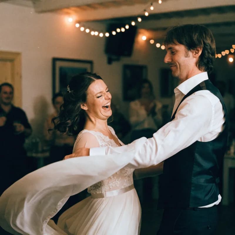 A bride mid turn on the dance floor of a string-lit function room, head tipped back laughing, her new husband watching her rather than the room.