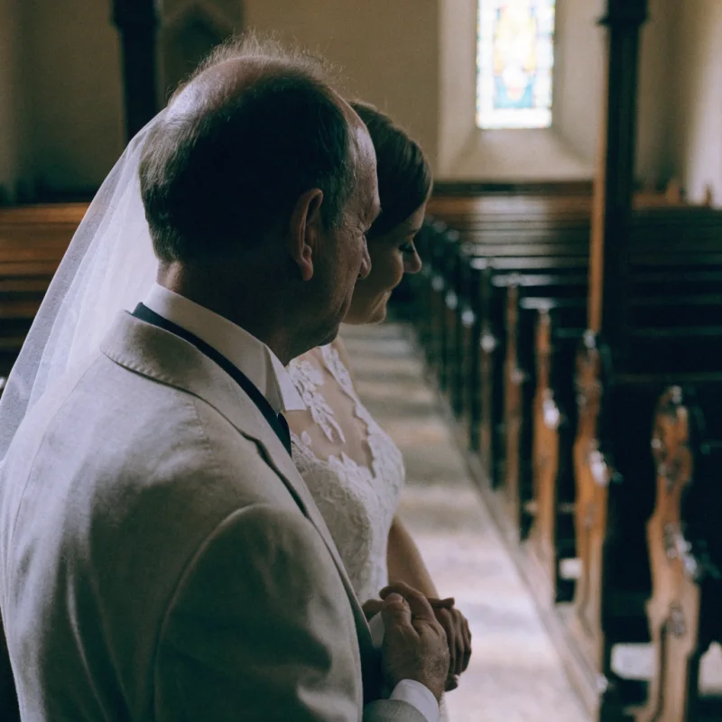 A father in a morning suit at the back of a village church with his daughter on his arm, both looking down the aisle.