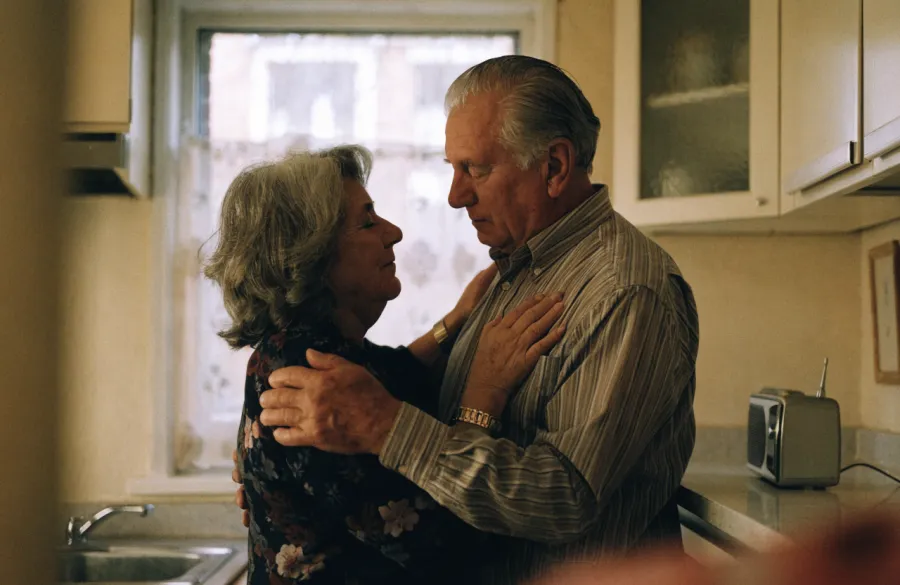 A married couple in their seventies holding each other and swaying to the radio in their kitchen.