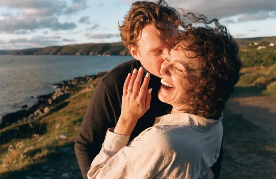 A couple embracing on a coastal path at golden hour, an engagement ring on her raised hand.