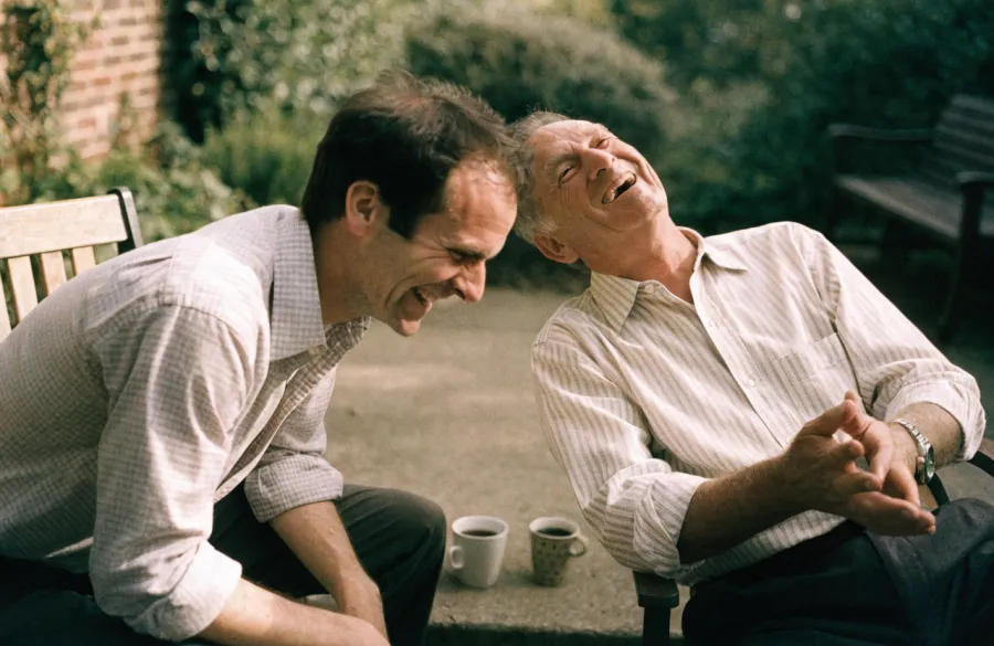 An adult son and his father laughing together on garden chairs, mugs of tea beside them.