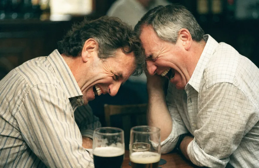 Two old friends in a pub leaning into each other, laughing helplessly.