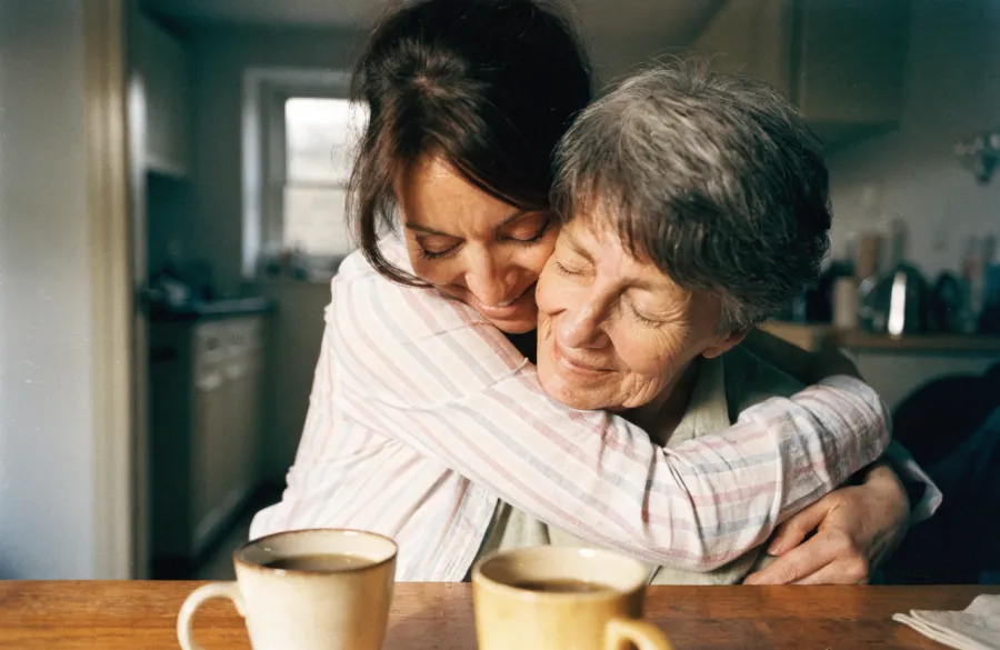 An adult daughter hugging her mother from behind at a kitchen table, both with their eyes closed.