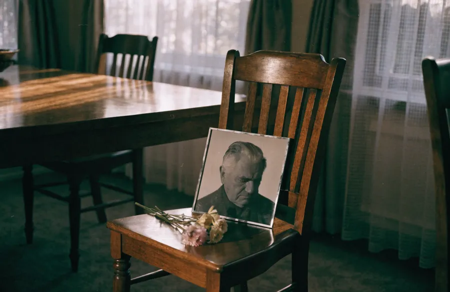 An empty dining chair with a framed black and white portrait of an older man resting on the seat beside a posy of flowers.