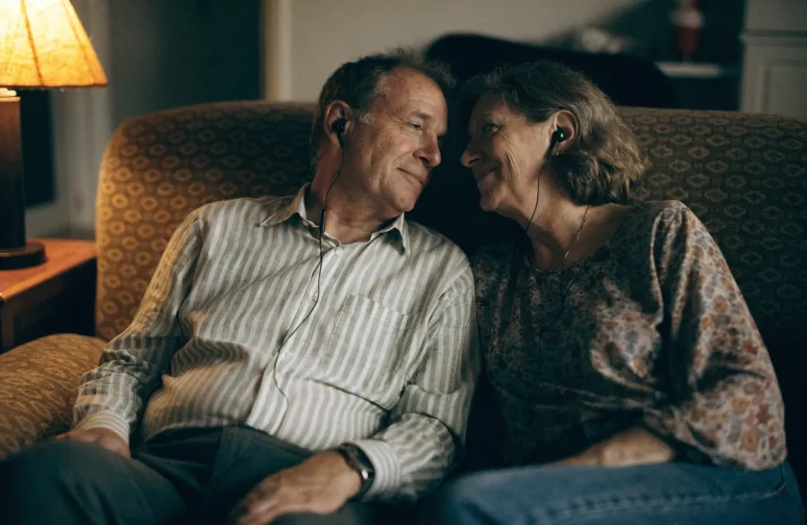 A couple in their fifties on a sofa sharing a pair of headphones, one earbud each.