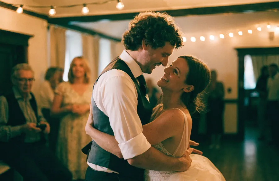 A bride and groom mid first dance, foreheads together, guests clapping around them.